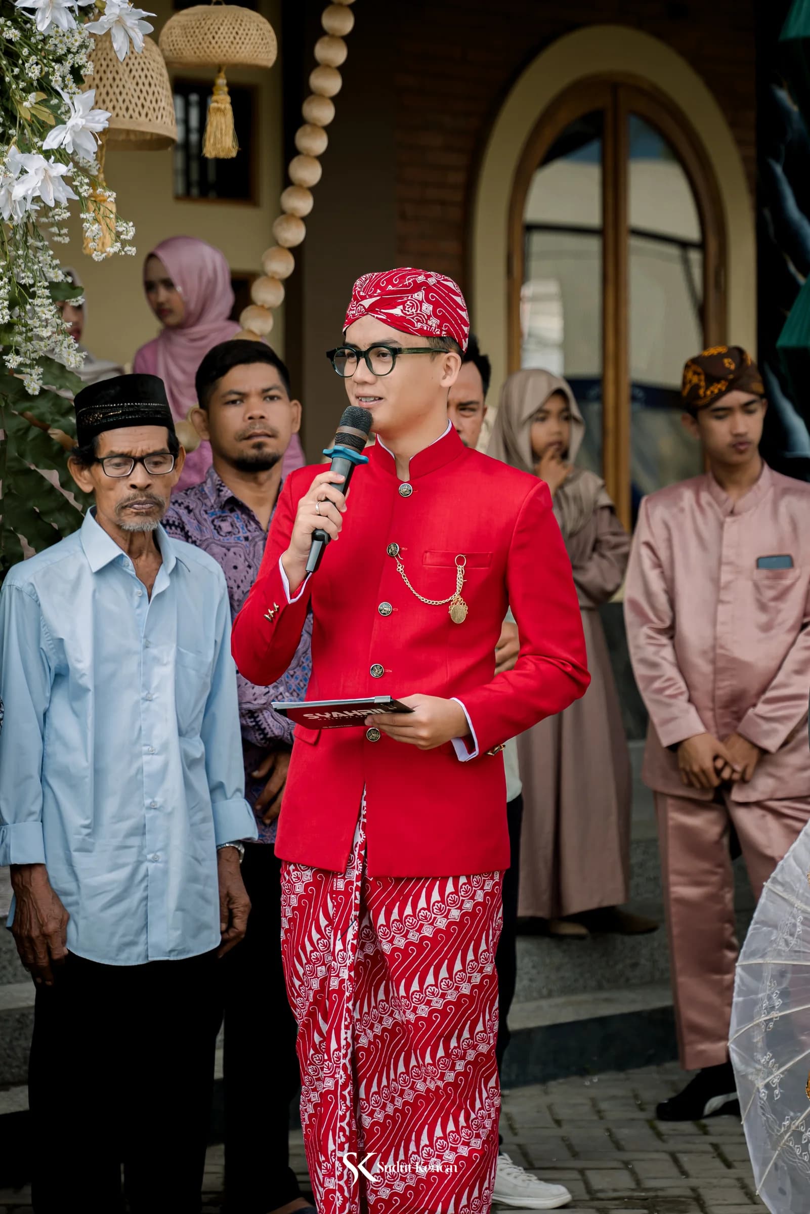 MC Syahril mengenakan beskap merah marun memandu kirab pengantin